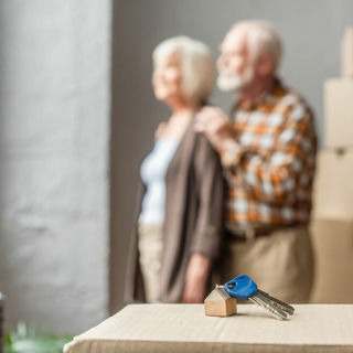 Twee oudere mensen staan uit het raam te kijken voor hen staat een verhuisdoos met sleutels daarop