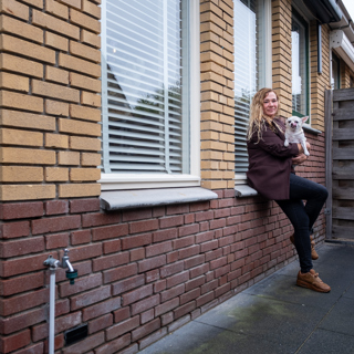 Vrouw zit met haar hond op schoot op de vensterbank voor het raam buiten haar woning.